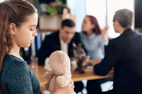 Young girl holding a teddy bear while parents argue with attorneys in the background during a child support and visitation dispute.